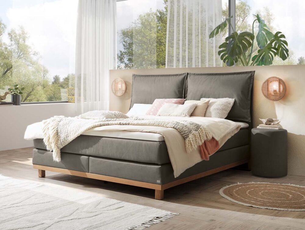 Ein modernes Schlafzimmer mit einem großen grauen Polsterbett, weißer und beiger Bettwäsche, Akzentkissen, weichen Überwürfen, einem runden Beistelltisch, einer Topfpflanze, geflochtenen Lampen, einem hellen Holzboden und großen Fenstern mit transparenten Vorhängen.