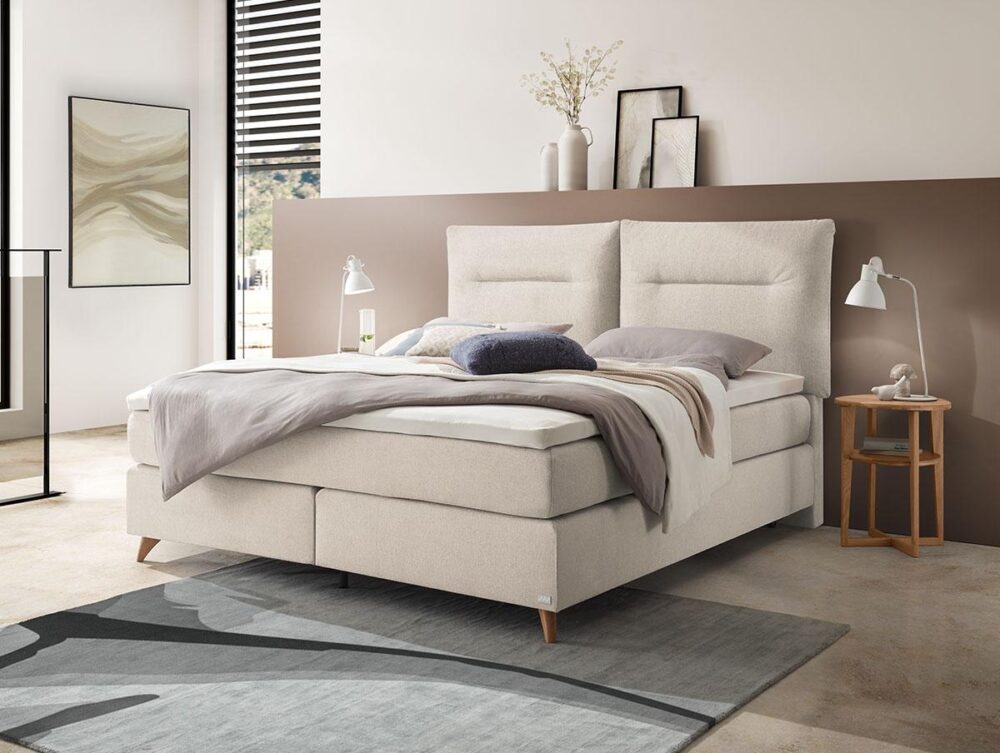 Modernes Schlafzimmer mit einem großen cremefarbenen Polsterbett, neutraler Bettwäsche, Nachttischen mit weißen Lampen, abstrakter Wandkunst und dekorativen Vasen. Sonnenlicht strömt durch ein Fenster mit schwarzen Jalousien.