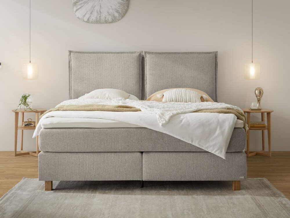 Ein modernes Schlafzimmer mit einem großen Polsterbett, neutraler Bettwäsche, zwei hölzernen Nachttischen mit Lampen und Dekorationsartikeln; ein weicher Vorleger und Pendelleuchten schaffen eine gemütliche Atmosphäre.