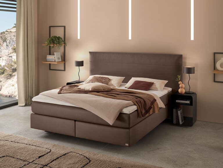 Ein modernes Schlafzimmer mit einem großen braunen Polsterbett, beiger Bettwäsche, zwei schwarzen Nachttischen mit Lampen, Regalen mit Dekoration und raumhohen Fenstern mit Blick auf die Felsen draußen.