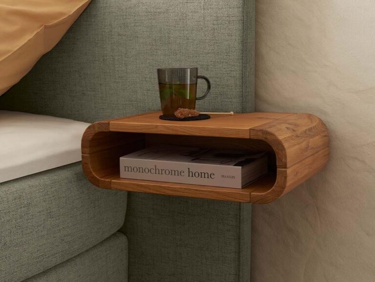 Ein schwebender Nachttisch aus Holz, der neben einem Bett an der Wand befestigt ist und auf dem ein Glas mit einem Getränk und einem Keks steht, während im Regal darunter ein Buch mit dem Titel Monochrome Home liegt.