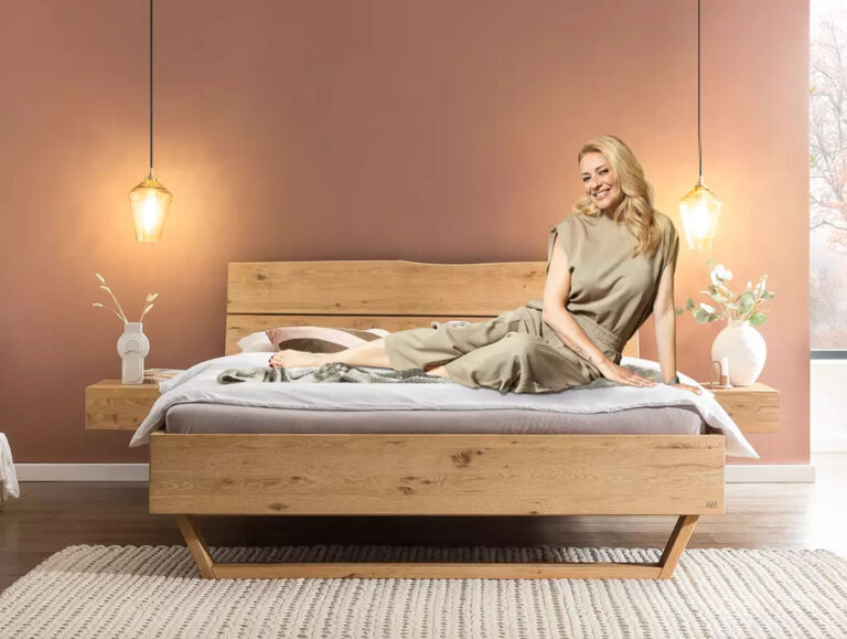 Eine lächelnde Frau in einem beigen Outfit sitzt auf einem modernen Holzbett mit einer beigen Decke in einem gemütlichen Schlafzimmer. Prominente Empfehlung: Christa Rigozzi setzt auf Bettenland für stilvolles Design und Komfort.
