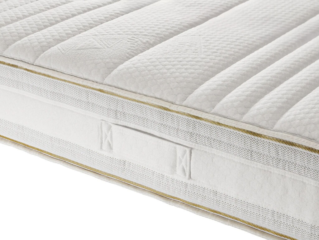 Die Nahaufnahme der Talalay-Latexmatratzenhülle "DreamOn" zeigt ihre gesteppte, strukturierte Oberfläche, den goldenen Reißverschluss und den seitlichen Stoffgriff. Die Matratze hat horizontale Steppnähte und ein dickes, plüschiges Aussehen. Sie ist mit Schafschurwolle gefüllt.