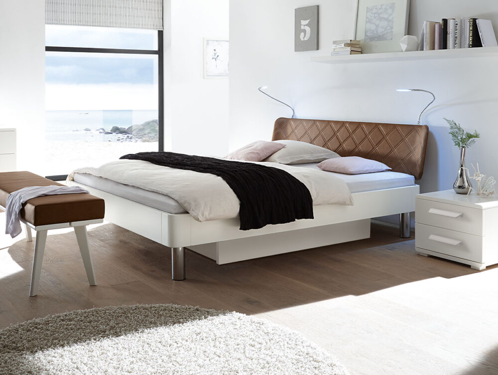 Ein modernes, sonnendurchflutetes Schlafzimmer mit einem weißen Bett mit einem braunen getufteten Kopfteil, weißer Bettwäsche und einer dunklen Decke. Es gibt einen Nachttisch mit einer Vase, eine Bank, Wandregale mit Dekoration und ein großes Fenster mit Blick auf den Strand.