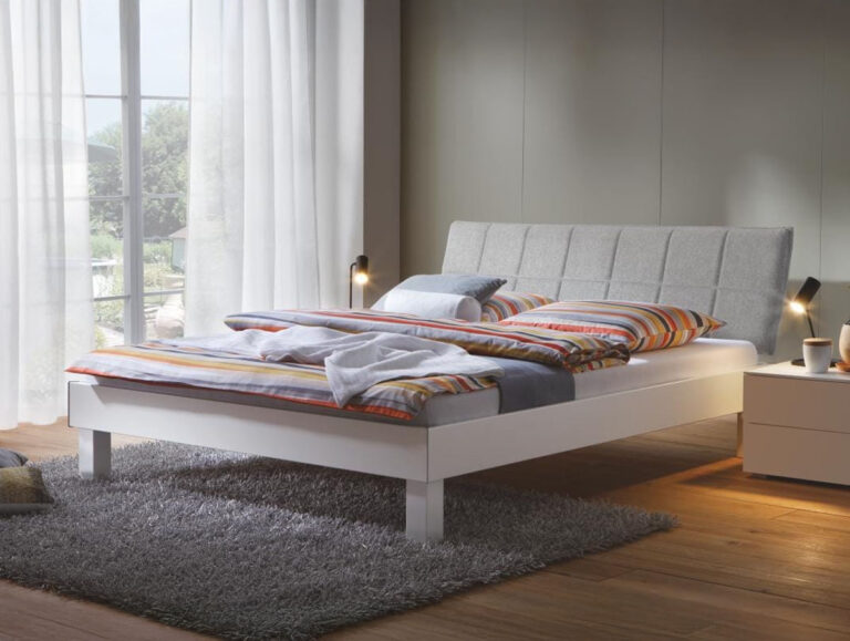 Ein modernes Schlafzimmer mit einem grauen Polsterbett, bunt gestreifter Bettwäsche, zwei Nachttischen mit kleinen Lampen, einem grauen Teppich, einem Holzboden und großen Fenstern mit weißen Vorhängen, die das natürliche Licht hereinlassen.