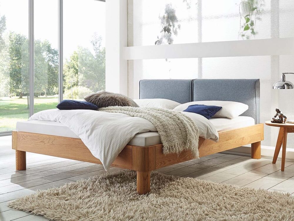 Ein modernes Schlafzimmer mit einem Bettgestell aus Holz, einem grau gepolsterten Kopfteil, weißer Bettwäsche und blauen Kissen. Eine kuschelige beige Decke und ein weicher Zottelteppich liegen auf dem Boden. Große Fenster bieten einen Blick auf das Grün draußen.