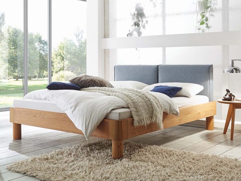 Ein modernes Schlafzimmer mit einem Bettgestell aus Holz, einem grau gepolsterten Kopfteil, weißer Bettwäsche und blauen Kissen. Eine kuschelige beige Decke und ein weicher Zottelteppich liegen auf dem Boden. Große Fenster bieten einen Blick auf das Grün draußen.