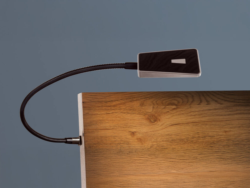 Eine biegsame, moderne Leselampe mit einem rechteckigen schwarzen Kopf ist an der Kante einer hölzernen Oberfläche vor einem schlichten grauen Hintergrund angebracht.