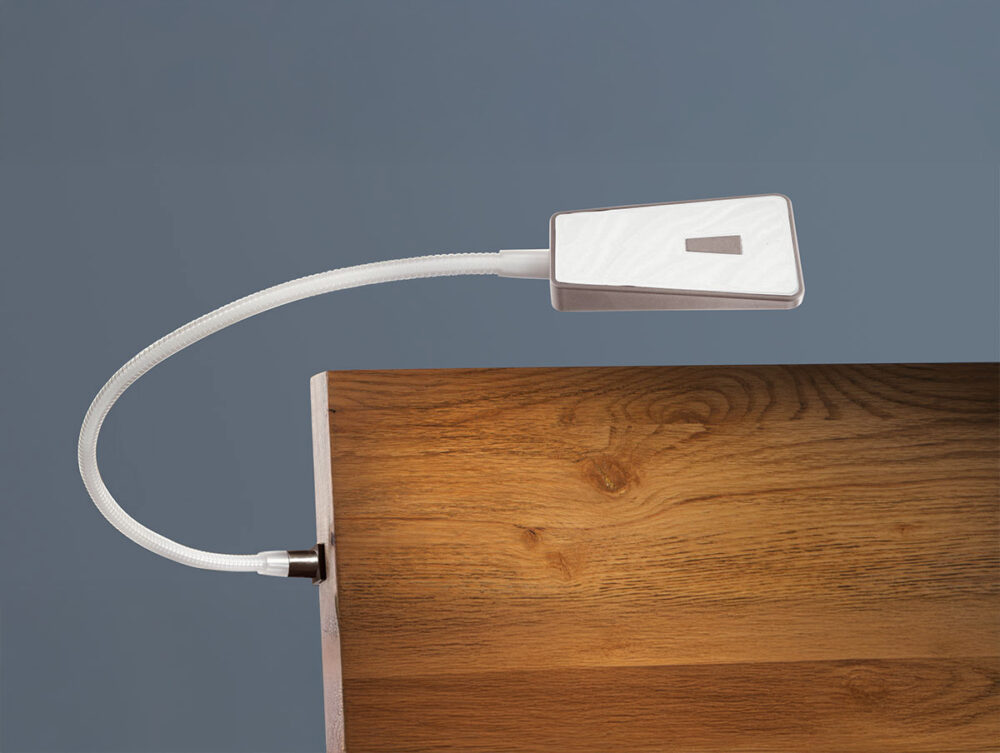 Eine biegsame Leselampe mit rechteckigem Kopf ist seitlich an einer hölzernen Oberfläche, wahrscheinlich einem Schreibtisch oder Tisch, vor einem schlichten grauen Hintergrund angebracht.