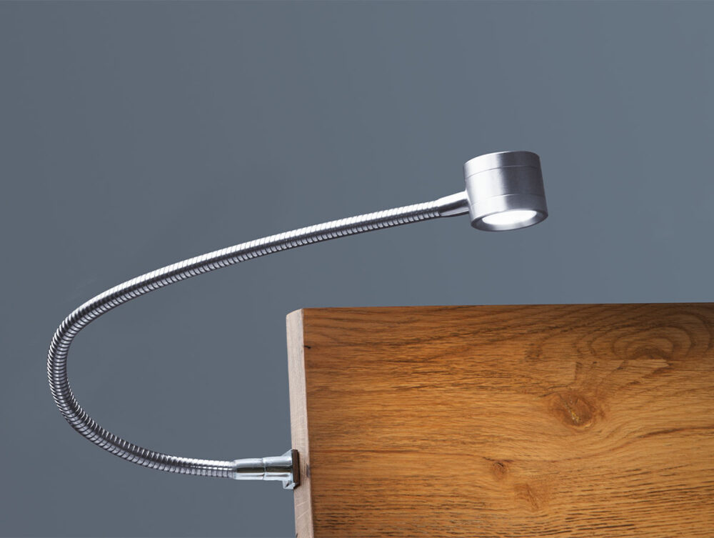 An der Seite eines hölzernen Kopfteils ist eine silberne, biegsame Leselampe angebracht, deren Licht nach unten auf einen schlichten grauen Hintergrund gerichtet ist.