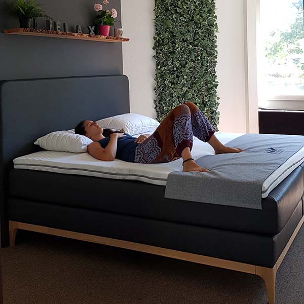 Eine Person liegt auf dem Rücken auf einem ordentlich gemachten Bett in einem hellen, modernen Schlafzimmer mit einem RELAX-Schild und einer Topfpflanze und genießt die Ruhe nach ihrer Guten Morgen Yoga - Morgenroutine zum Erwachen, mit vertikaler grüner Pflanzendekoration an der Wand.