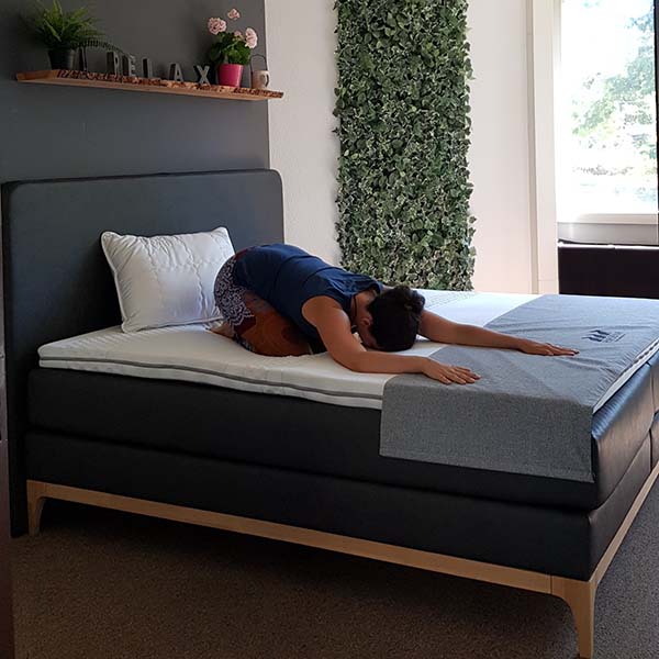 Eine Person kniet in kindlicher Haltung auf einem Bett und streckt sich mit ausgebreiteten Armen nach vorne. Der Raum hat ein graues Kopfteil, weißes Bettzeug, Pflanzen und Sonnenlicht, das durch ein Fenster einfällt.