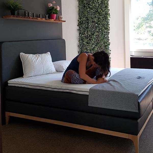 Eine Frau sitzt im Schneidersitz auf einem ordentlich gemachten Bett und stützt ihren Kopf entspannt auf ihre Knie. Der Raum hat eine grüne Wanddekoration, ein Regal mit Dekoration und Sonnenlicht, das in den Raum fällt.