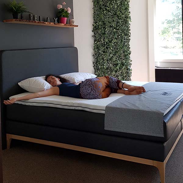 Eine Frau liegt auf dem Rücken mit ausgestreckten Armen und einem Bein auf einem sauber gemachten Bett in einem modernen, hellen Raum mit einem RELAX-Schild und Pflanzen an der Wand.