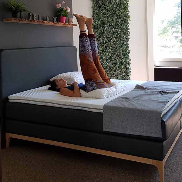 Eine Frau liegt auf einem Bett und stützt sich mit hochgelegten Beinen auf dem Kopfteil ab. Sie trägt eine gemusterte Hose und ein dunkles Oberteil. Der Raum ist modern gestaltet, mit grünem Blattdekor an der Wand und einem RELAX-Schild auf einem Regal.