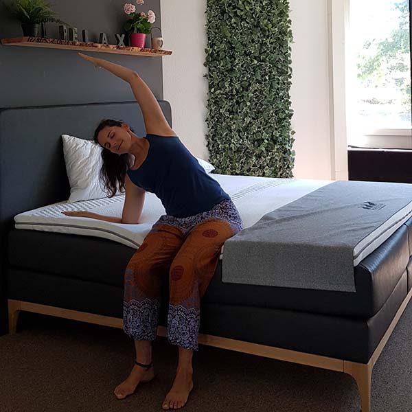 Eine Frau sitzt auf der Kante eines Bettes und streckt ihren linken Arm über den Kopf, während ihr rechter Arm auf der Matratze ruht. Sie trägt ein marineblaues Tank-Top und eine gemusterte Hose. Die Wand ist begrünt und das Fenster ist sonnendurchflutet.