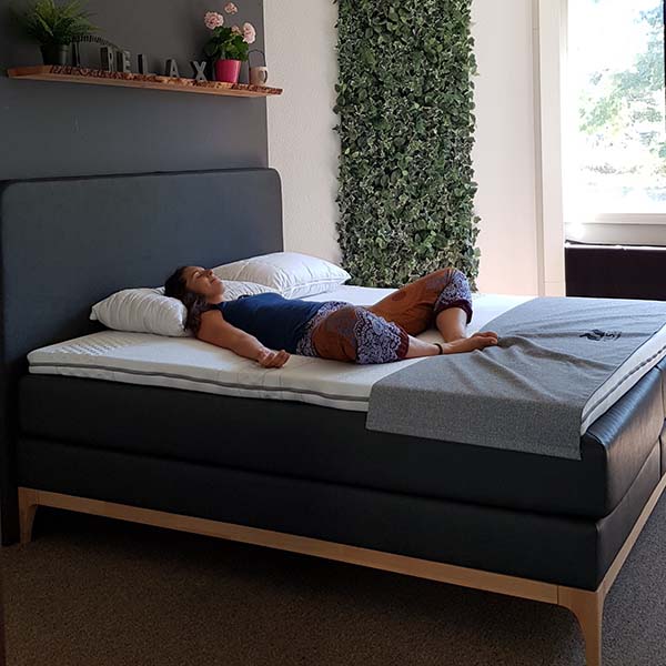 In einem hellen Raum liegt eine Frau mit ausgebreiteten Armen und geschlossenen Augen auf dem Rücken auf einem ordentlich gemachten Bett. Hinter ihr steht ein Regal mit dem Wort RELAX und Pflanzen, und eine vertikale grüne Pflanzenwand schmückt den Raum.