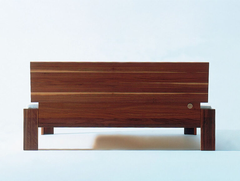 Eine minimalistische Holzbank mit einer massiven, rechteckigen Rückenlehne und Sitzfläche, die sich durch klare Linien und eine warme, braune Holzoberfläche vor einem schlichten, hellen Hintergrund auszeichnet.