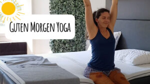 Eine Frau sitzt auf ihrem Bett in einer Yogapose, streckt ihre Arme nach oben und lächelt. Der Raum ist hell, mit einer Sonnengrafik und dem Text Guten Morgen Yoga auf Deutsch auf der linken Seite.