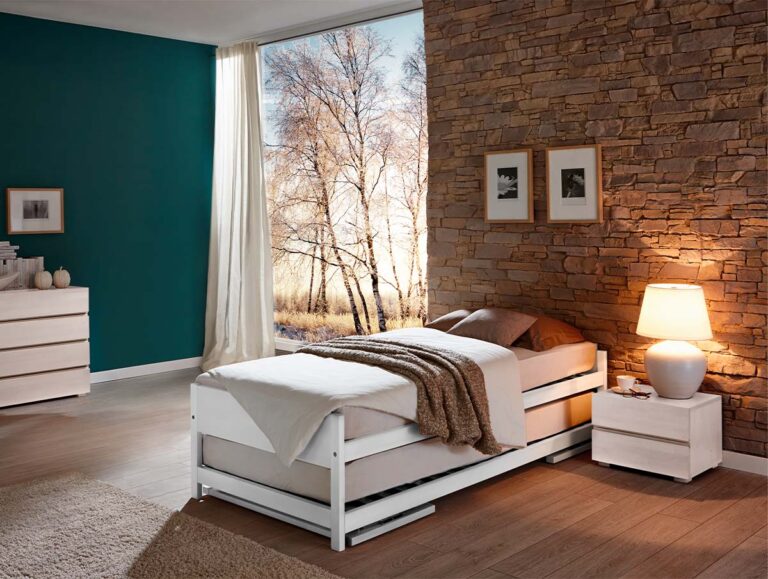 Ein gemütliches Schlafzimmer mit einem Hasena Gästebett "Lido" in brauner Bettwäsche, neben einer brennenden Lampe auf einem Nachttisch, einer Akzentwand aus Stein, gerahmten Bildern, einer Kommode und einem großen Fenster mit kahlen Bäumen draußen.