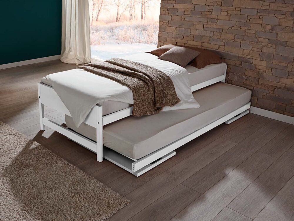 Ein weißes Hasena Gästebett "Lido" mit Bettzeug und Kissen steht auf einem Holzboden an einer Steinmauer, die durch das Fenster in Sonnenlicht getaucht wird und einen ruhigen Blick auf die Bäume draußen bietet.