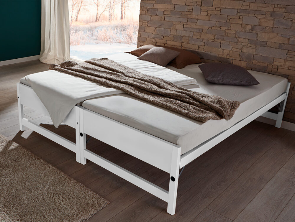 Das Hasena Gästebett "Lido" mit weißem Gestell, beiger Bettwäsche, braunen Kissen und einer strukturierten Decke steht in einem Raum mit einer Akzentwand aus Stein und einem großen Fenster mit winterlicher Aussicht.