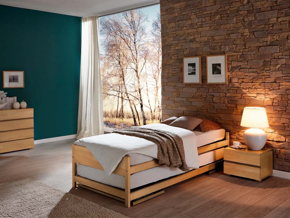 Ein gemütliches Schlafzimmer mit einem Hasena Gästebett "Lido", weißer Bettwäsche, einer braunen Überwurfdecke, einer Akzentwand aus Stein mit gerahmten Fotos, einem Nachttisch mit Lampe, einer Kommode und einem großen Fenster mit Blick auf die sonnenbeschienenen Bäume draußen.