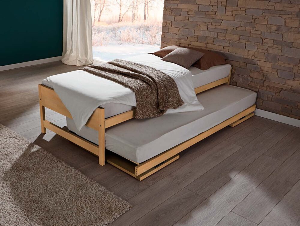 Das Hasena Gästebett "Lido" mit weißer Bettwäsche und braunen Decken steht in einem gemütlichen Zimmer mit Holzböden, einer Akzentwand aus Stein und einem großen Fenster mit Blick auf eine verschneite Landschaft.