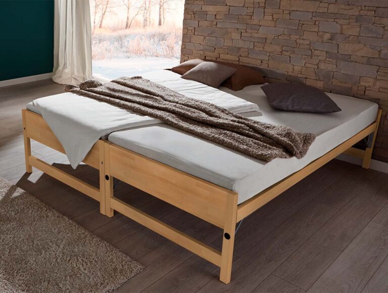 In einem gemütlichen, sonnendurchfluteten Schlafzimmer mit Holzfußboden steht das Hasena Gästebett "Lido", das mit weißer Bettwäsche, braunen und grauen Kissen und einer braunen Decke ausgestattet ist und an einer Steinwand steht.