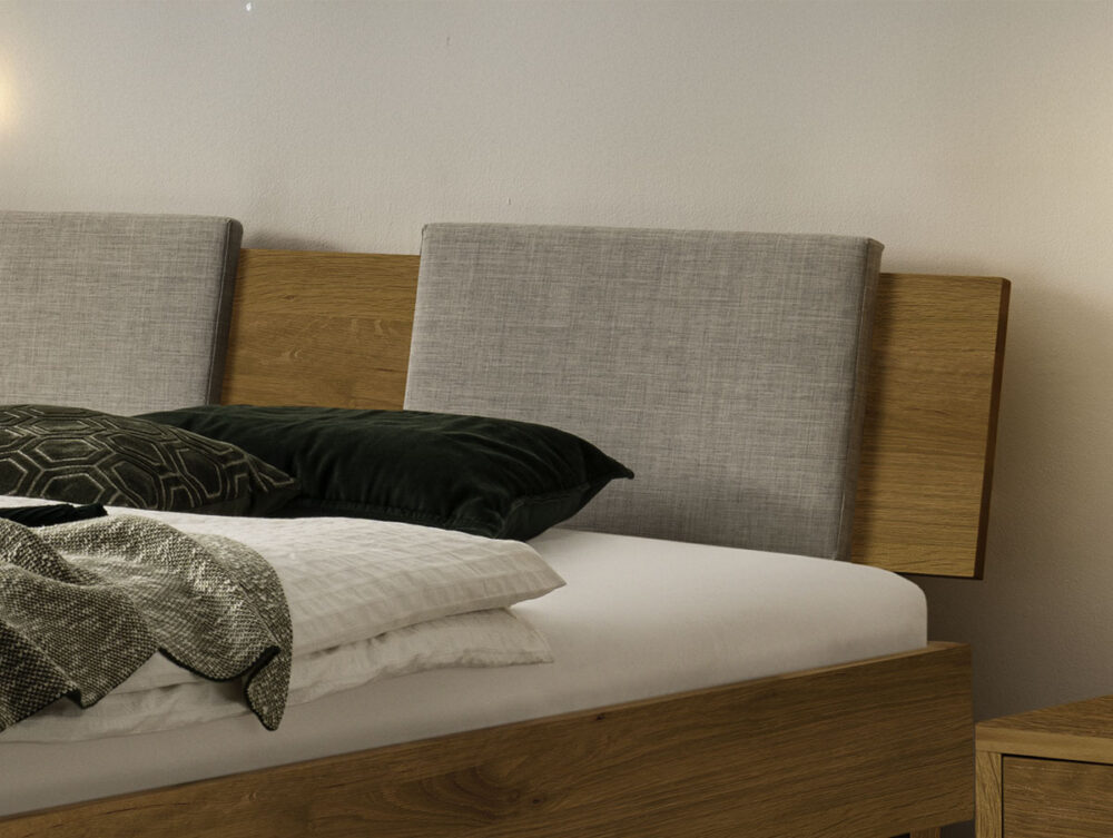 Nahaufnahme eines modernen Holzbettes mit einem grau gepolsterten Kopfteil, weißer Bettwäsche, einem grünen Überwurfkissen und einem geometrisch gemusterten Kissen neben einem passenden Holznachttisch.