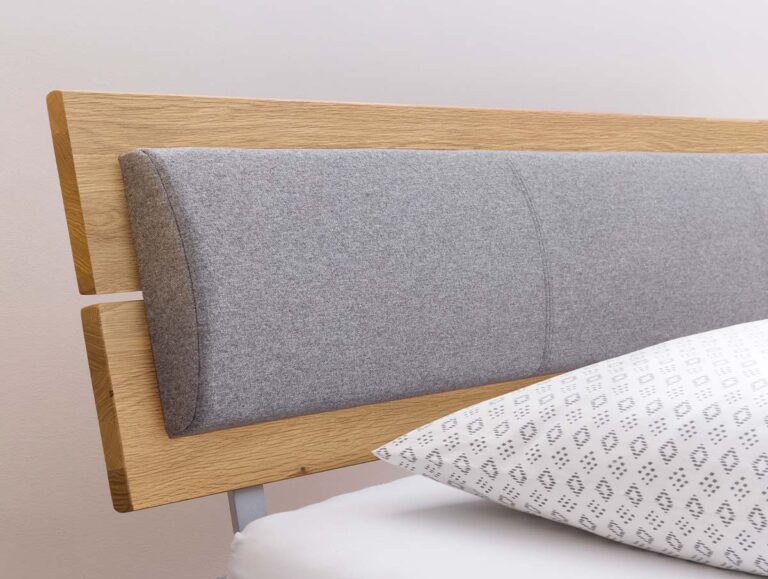 Nahaufnahme eines modernen Holzbettes mit einem hellgrau gepolsterten Kopfteil, einem weißen Kissen mit geometrischem Muster und einer Topfpflanze auf einem Tisch in der Nähe.