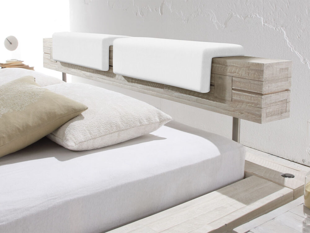 Ein modernes Bett mit einem rustikalen Holzkopfteil und Hasena Kopfteilkissen "Nakio" wird mit zwei weißen gefalteten Handtüchern bedeckt, mit weißer Bettwäsche und beigen Kissen bekleidet und vor einer strukturierten weißen Wand aufgestellt.