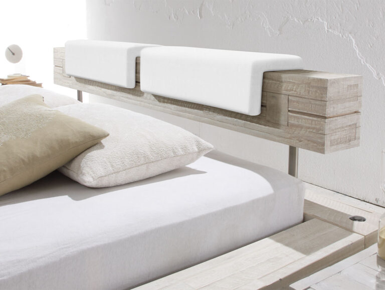 Ein modernes Bett mit einem rustikalen Holzkopfteil und Hasena Kopfteilkissen "Nakio" wird mit zwei weißen gefalteten Handtüchern bedeckt, mit weißer Bettwäsche und beigen Kissen bekleidet und vor einer strukturierten weißen Wand aufgestellt.