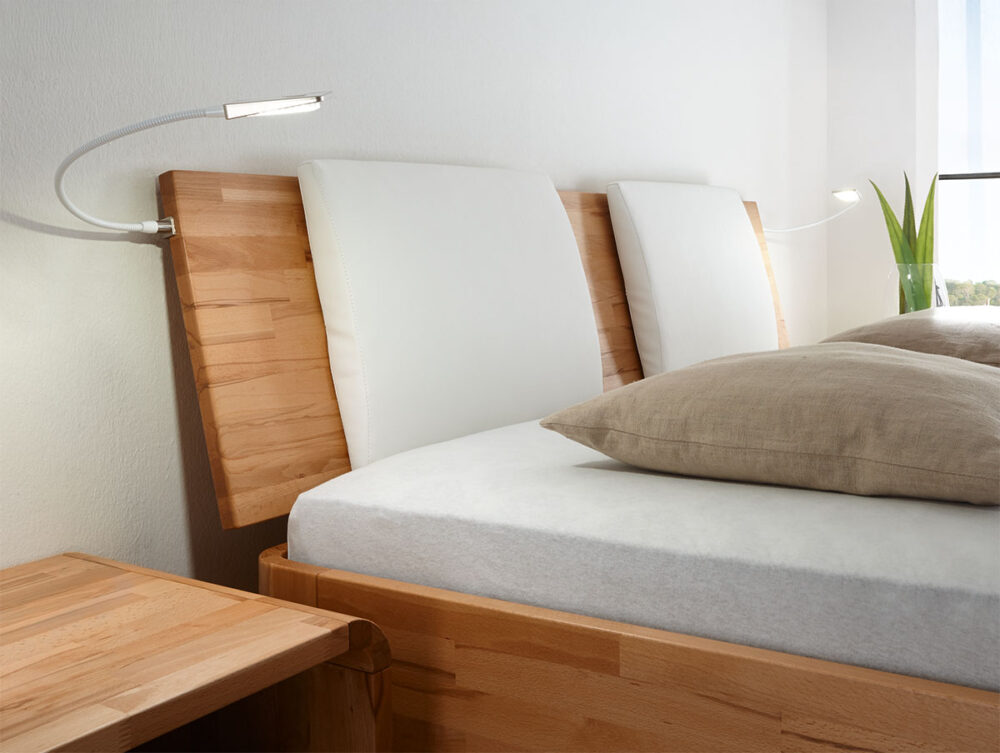 Ein modernes Holzbett mit einer weißen Matratze, beigen Kissen und weiß gepolsterten Kopfteilen. Am Kopfteil sind flexible Leselampen angebracht, und auf der Fensterbank im Hintergrund steht eine kleine Pflanze.
