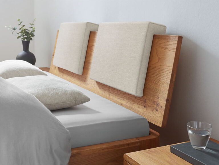 Ein Holzbett mit geneigtem Kopfteil und zwei quadratischen beigen Kissen, graues Bettzeug, zwei Kissen, ein Nachttisch mit einem Wasserglas und eine Vase mit grünen Zweigen im Hintergrund.