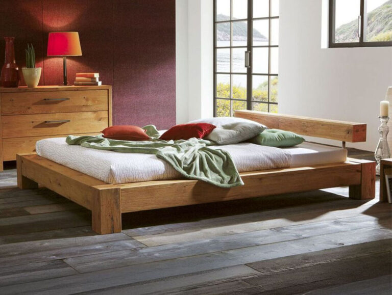 In einem stilvollen Schlafzimmer steht das Hasena Bett "Oak-Wild Bormio 1" aus Holz mit einer weißen Matratze, einer grünen Decke und bunten Kissen. In der Nähe steht eine Holzkommode mit einer roten Lampe, während große Fenster natürliches Licht und Grün hereinlassen.