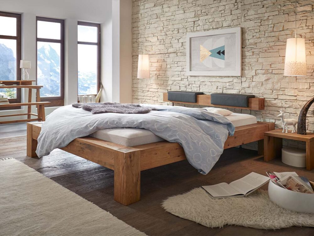 Ein gemütliches Schlafzimmer mit dem Hasena Bett "Oak-Wild Bormio 3", hellblauer Bettwäsche, einer Akzentwand aus Stein und großen Fenstern mit Bergblick. Ein weicher Teppich, ein offenes Buch und dekorative Akzente bringen Wärme in den modernen, rustikalen Raum.