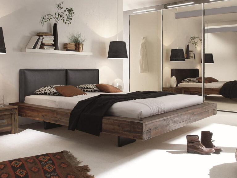 Ein modernes Schlafzimmer mit dem Hasena Bett "Factory-Line Bloc Vilo" Holzbett, schwarzem Kopfteil, weißer Bettwäsche, braunen und schwarzen Kissen, verspiegelten Schranktüren, Hängelampen, einem Regal mit Büchern und Pflanzen und braunen Stiefeln auf dem Boden.