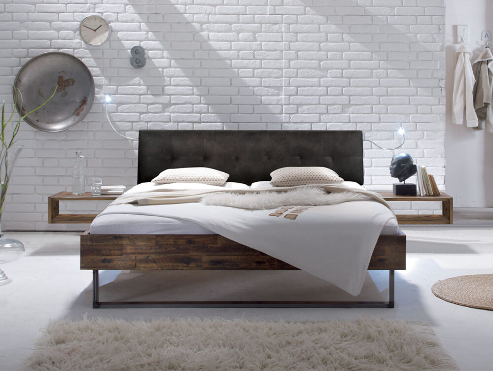 Ein modernes Schlafzimmer präsentiert das Hasena Bett "Factory-Line Loft Indus" mit einem dunklen Kopfteil, weißem Bettzeug, hölzernen Nachttischen, einer weißen Ziegelwand, dekorativen Akzenten und einem beigen Teppich für eine gemütliche, minimalistische Atmosphäre.