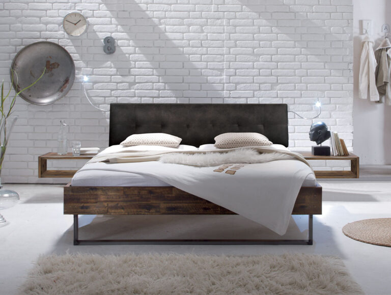 Ein modernes Schlafzimmer präsentiert das Hasena Bett "Factory-Line Loft Indus" mit einem dunklen Kopfteil, weißem Bettzeug, hölzernen Nachttischen, einer weißen Ziegelwand, dekorativen Akzenten und einem beigen Teppich für eine gemütliche, minimalistische Atmosphäre.