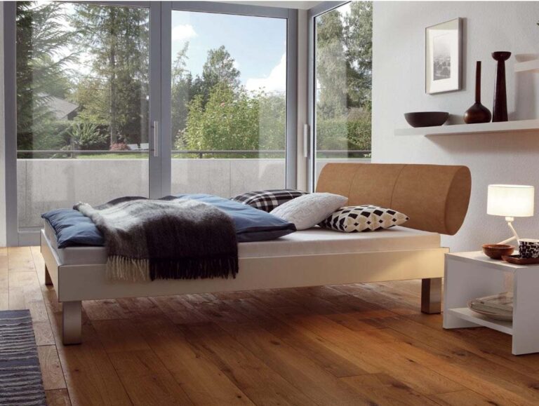 Ein modernes Schlafzimmer präsentiert das Hasena Bett "Movie-Line Star Mico 1" mit einem braunen Kopfteil, blaugrauer Bettwäsche, Holzboden und minimalistischer Wandkunst. Große Fenster geben den Blick auf Bäume und einen Balkon frei; Regale mit Dekoration sorgen für einen gemütlichen Touch.