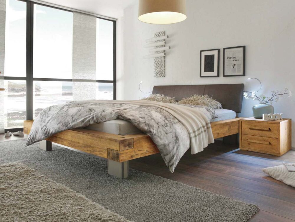 Modernes Schlafzimmer mit dem Hasena Bett "Oak-Vintage Bloc Quada" Holzbettgestell, grauer Bettwäsche und Plüschkissen. Große Fenster erhellen den Raum, der durch einen Nachttisch, einen gemütlichen Teppich und gerahmte Kunstwerke stilvoll ergänzt wird.