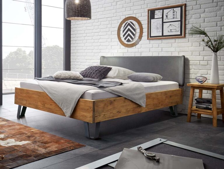 Ein modernes Schlafzimmer mit einem Bettgestell aus Holz, einem grauen Kopfteil und neutraler Bettwäsche. Das Zimmer hat weiße Backsteinwände, große Fenster, Wandkunst, einen hölzernen Beistelltisch mit Dekoration und eine hängende geflochtene Lampe.