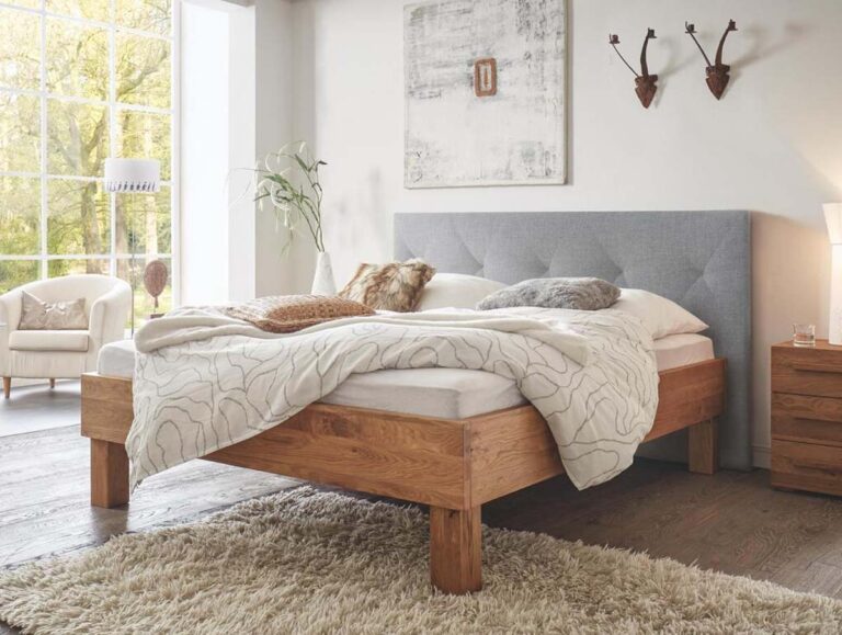 Ein modernes Schlafzimmer mit einem Hasena Bett "Oak-Wild Cadro 23 Ivio" Bettgestell, grauem Polsterkopfteil, heller Bettwäsche und gemütlichen Kissen. Der Raum verfügt über ein großes Fenster, einen Sessel, Wandkunst, einen beigen Teppich und einen Holzboden.
