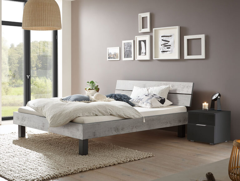 Modernes Schlafzimmer mit dem Hasena Bett "Top-Line Advance Mico 1", kuscheliger Bettwäsche und einem beigen Teppich. Eine Kommode, Wandkunst, große Fenster mit natürlichem Licht sowie gewebte und hölzerne Deko-Akzente schaffen eine helle, einladende Atmosphäre.