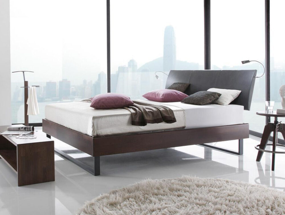 Ein modernes Schlafzimmer mit dem Hasena Bett "Wood-Line Premium 18 Indus", einer weißen Matratze und lila Kissen. Große Fenster bieten einen Blick auf die Skyline der Stadt, minimale Möbel sorgen für einen klaren Look, und ein runder beiger Teppich ergänzt den glänzenden Boden.