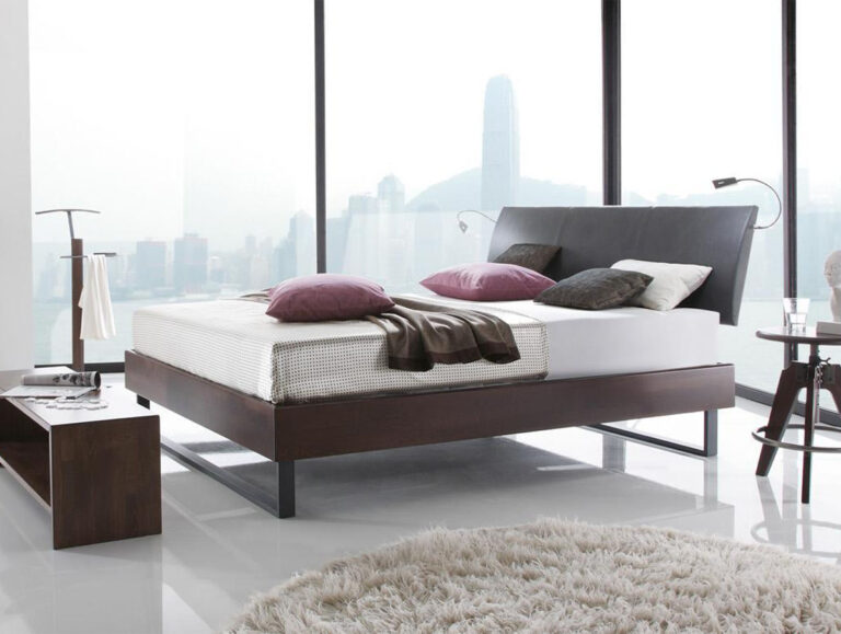 Ein modernes Schlafzimmer mit dem Hasena Bett "Wood-Line Premium 18 Indus", einer weißen Matratze und lila Kissen. Große Fenster bieten einen Blick auf die Skyline der Stadt, minimale Möbel sorgen für einen klaren Look, und ein runder beiger Teppich ergänzt den glänzenden Boden.