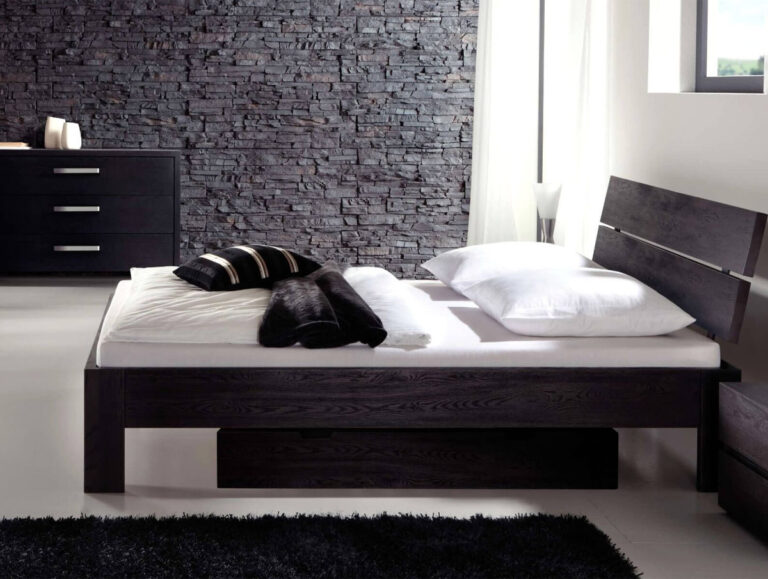 Ein modernes Schlafzimmer mit dem Hasena Bett "Oak-Line Modul Corno 1" aus dunklem Holz, weißem Bettzeug, schwarzen und weißen Kissen, einer Akzentwand aus Stein, einer schwarzen Kommode und großen Fenstern, die den Raum mit natürlichem Licht füllen.