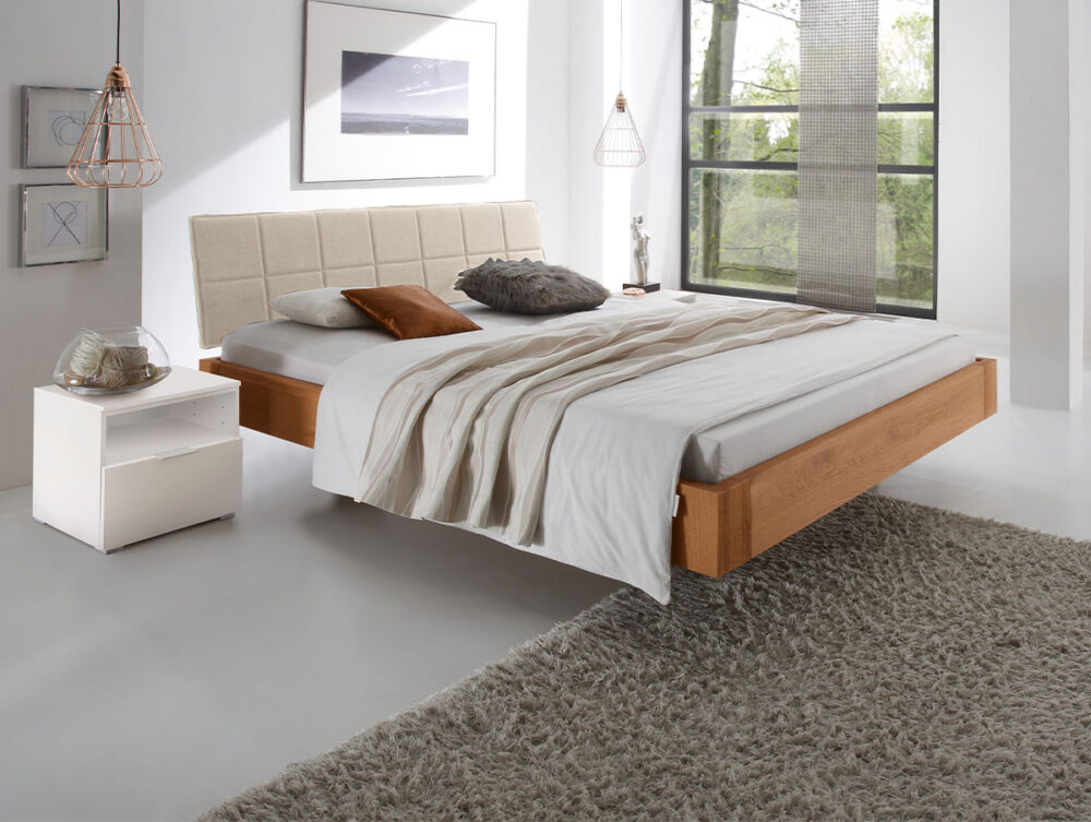 Ein modernes Schlafzimmer mit dem Hasena Bett "Oak-Line Modul Cora", einem beigefarbenen Polsterkopfteil, neutraler Bettwäsche, weißem Nachttisch, Hängelampen, Wandkunst, grauem Zottelteppich und großen Fenstern, die natürliches Licht hereinlassen.