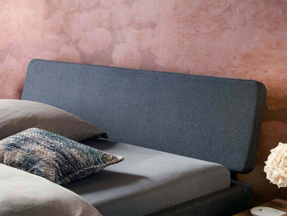 Ein modernes Bett mit einem dunkelgrau gepolsterten Kopfteil, grauer Bettwäsche, einem gemusterten Akzentkissen und einem Nachttisch mit einem Strauß weißer Blumen; die Wand dahinter ist mit einer zartrosa Blumentapete versehen.
